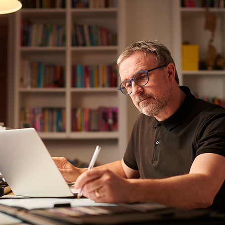 Mann mit Brille am Schreibtisch mit Laptop schaut auf Unterlagen